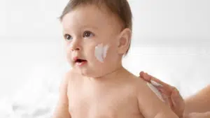Hydratation quotidienne : nourrisson recevant une application de crème hydratante. Application d'une crème hydratante sur le visage d'un nourrisson, illustrant l'importance des soins hydratants pour bébé.